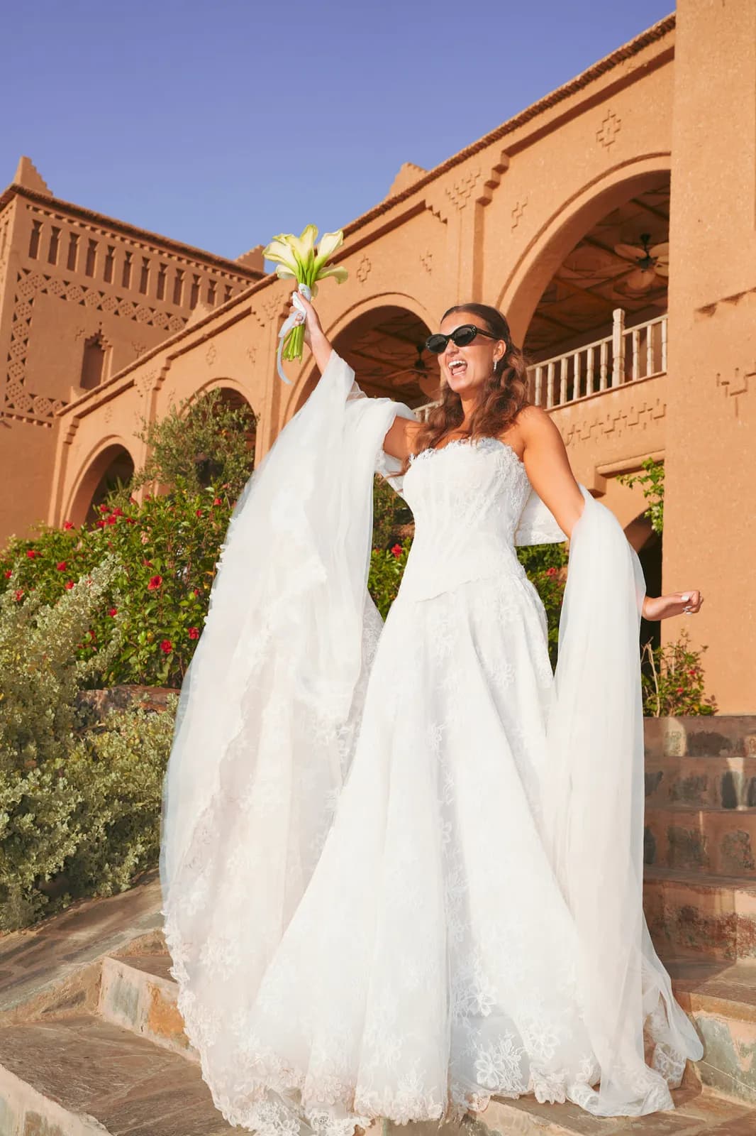 Bride laughing on the kasbah steps with her bouquet raised, Agafay desert wedding near Marrakech