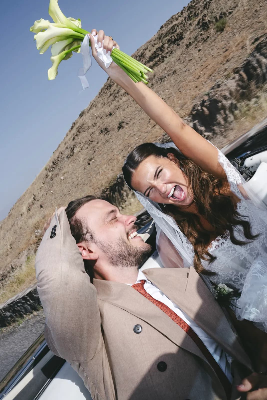 Bride cheering with her bouquet raised in a vintage car against the desert hills