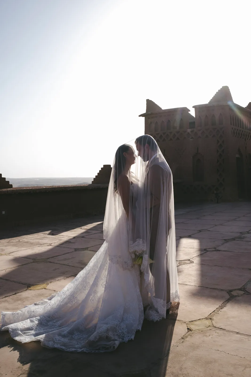 Bride against the carved earthen facade of an Agafay kasbah, black and white