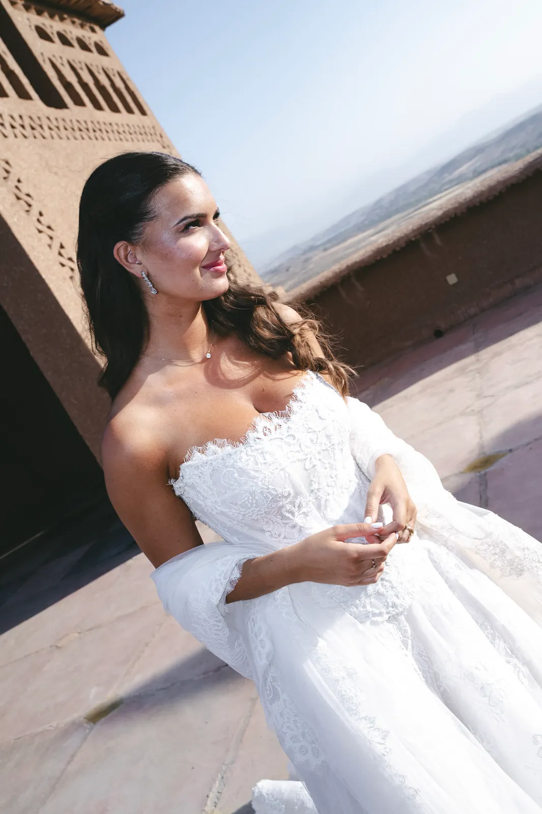 Bride in an off-shoulder lace gown seated by a kasbah tower above the desert